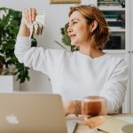 Smiling woman holding cash, working at home office with a laptop, symbolizing financial success.