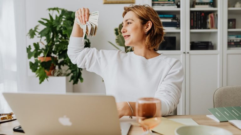 Smiling woman holding cash, working at home office with a laptop, symbolizing financial success.