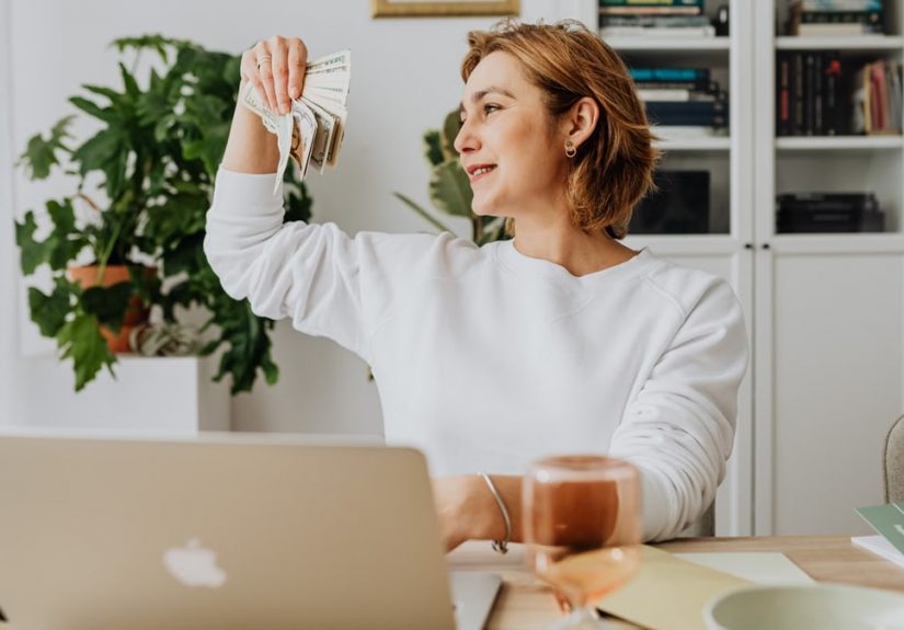 Smiling woman holding cash, working at home office with a laptop, symbolizing financial success.